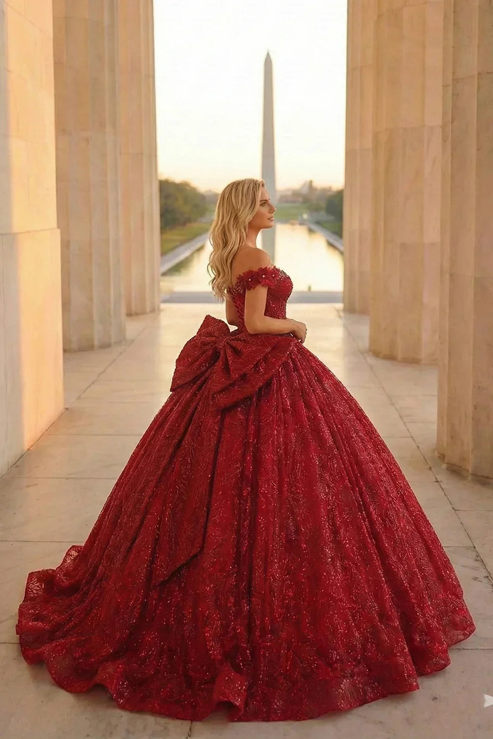 Woman in red ball gown with gold accents and large bow, standing outdoors at sunset