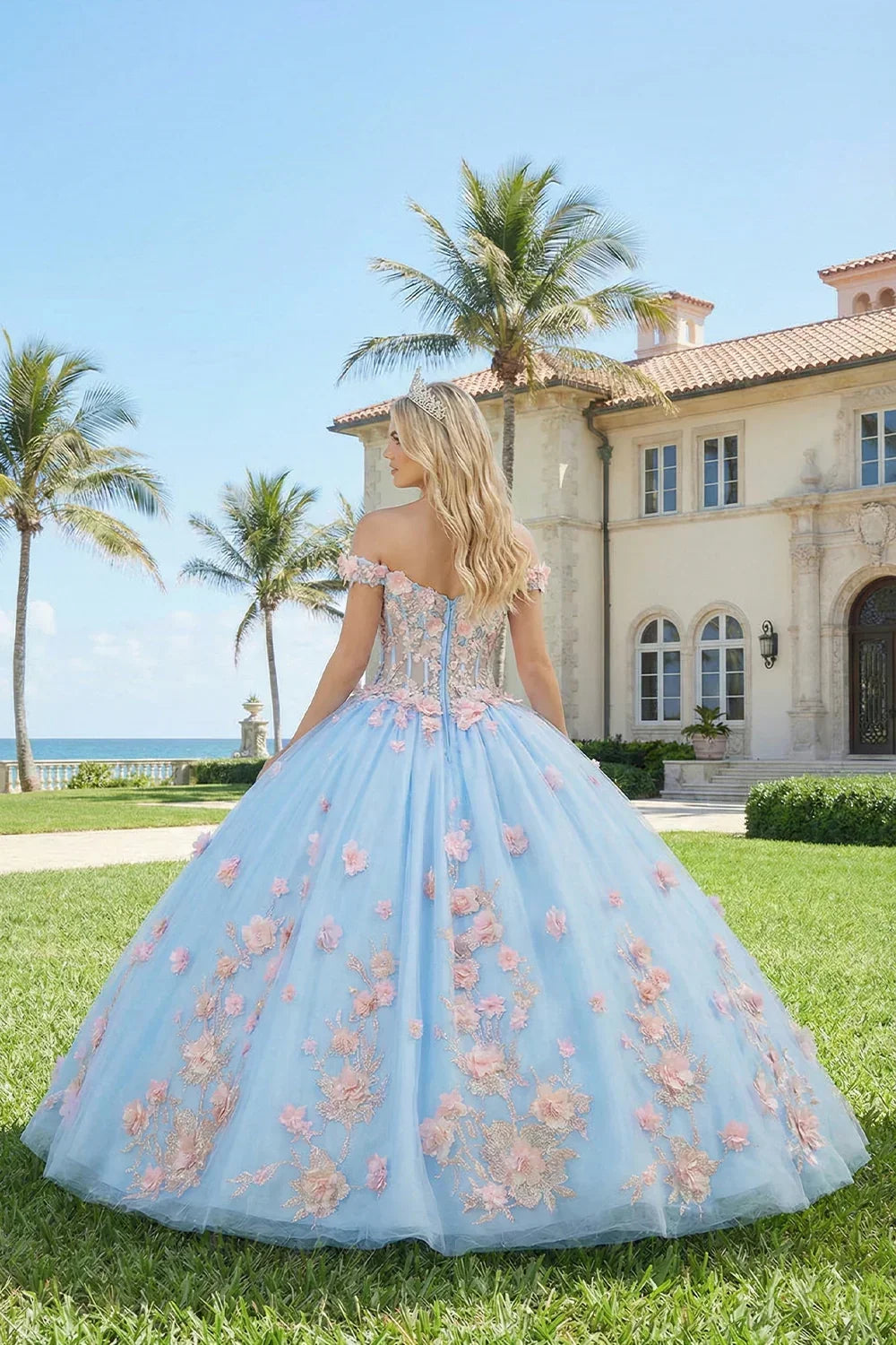 Woman in a blue ball gown with pink 3D floral lace outdoors by a mansion and palm trees