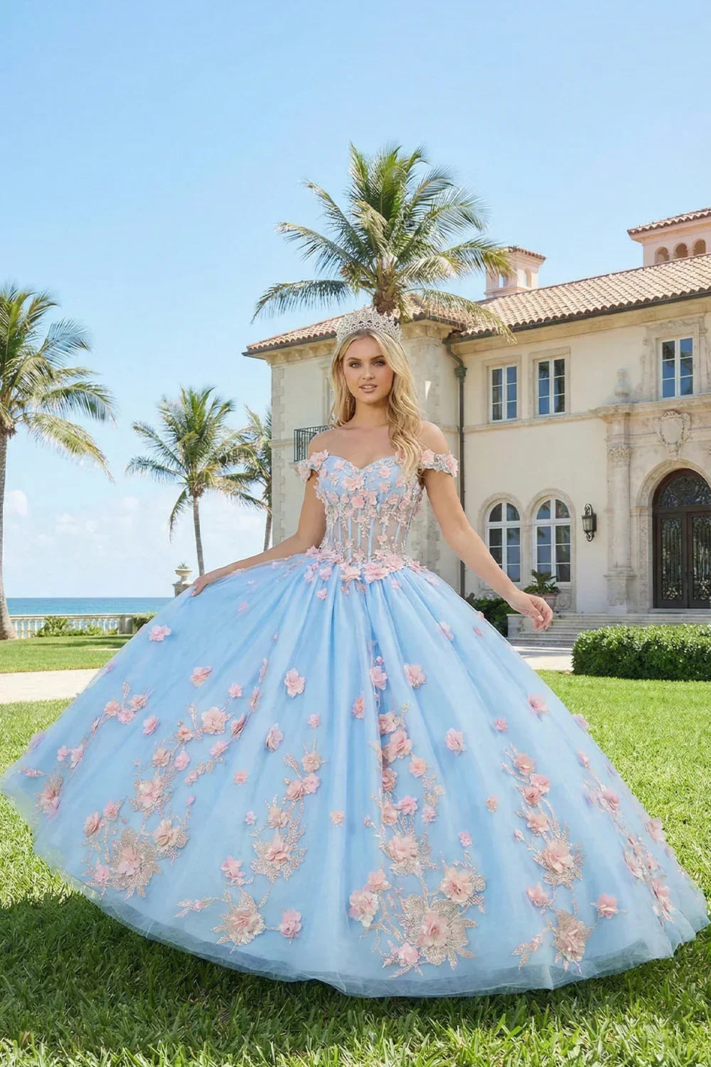 Woman in a blue ball gown with pink floral applique stands outdoors by a mansion