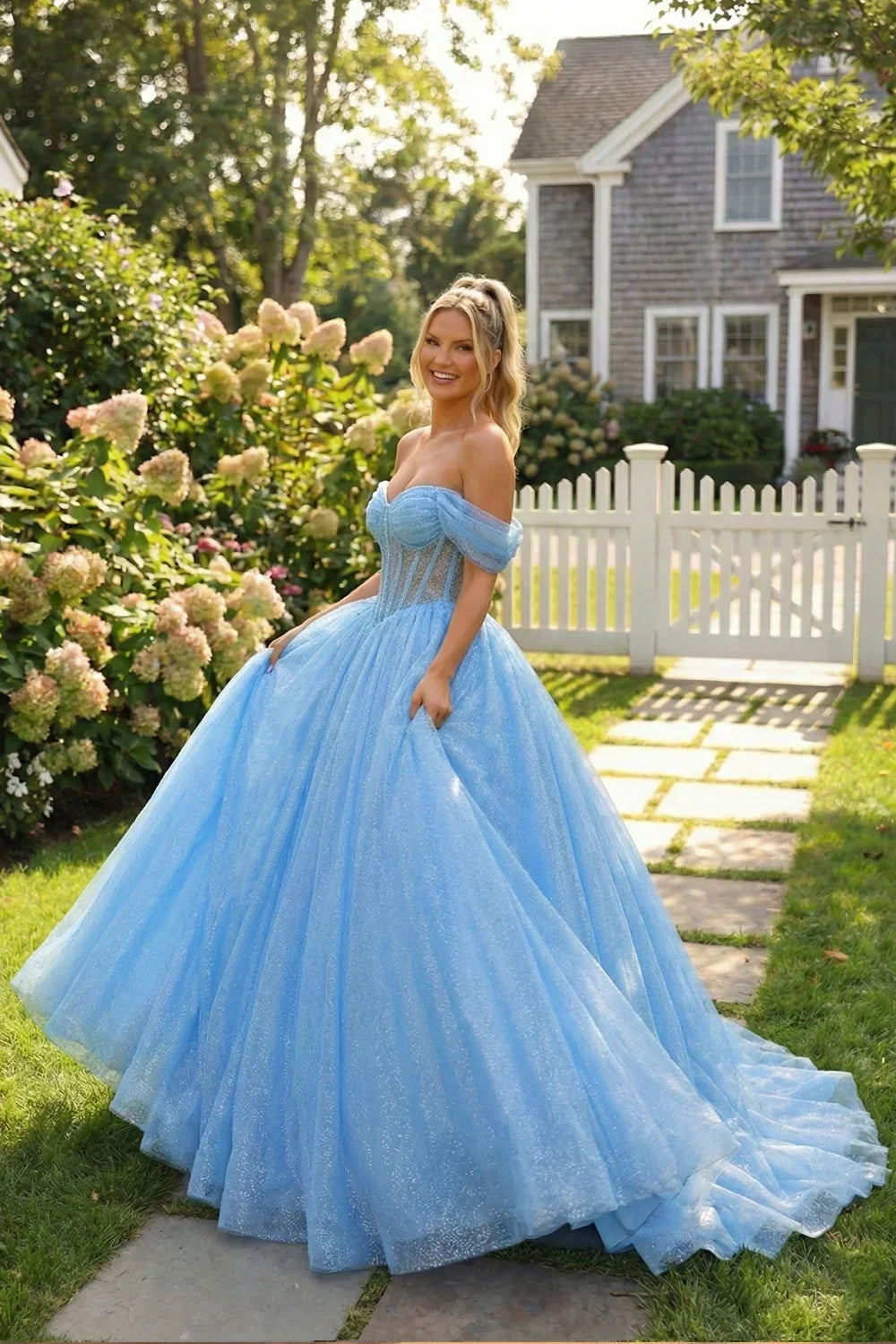 Woman in a sparkling blue ball gown dress with off-shoulder sleeves, standing outdoors by a white picket fence.