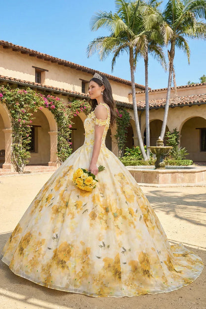 Woman in floral yellow ball gown holding yellow roses, wearing tiara, outdoors in courtyard